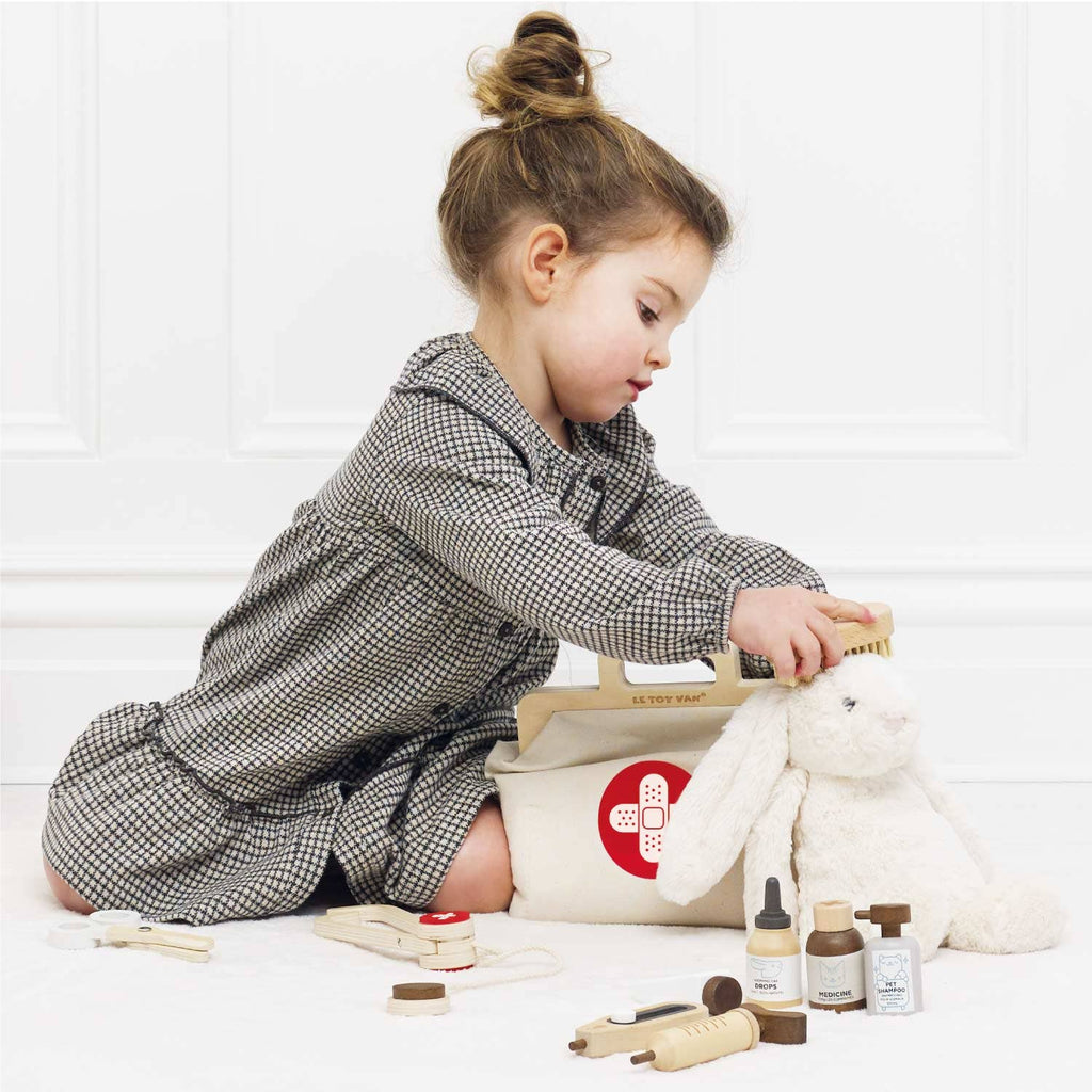A young girl in a checkered dress plays doctor with her Pretend Play Animal Vet Set, using toy medical tools and wooden veterinarian accessories to examine a white stuffed bunny while sitting on the floor beside a toy medical bag.