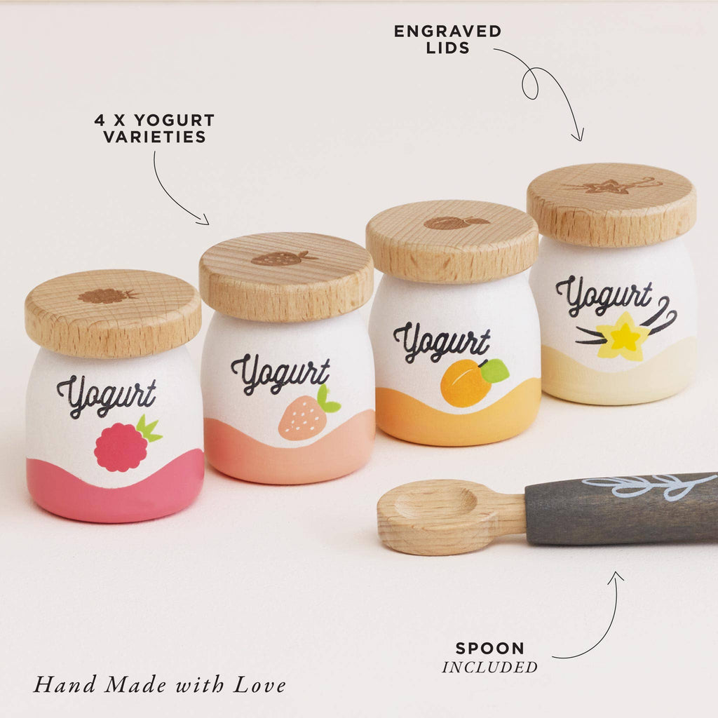 The Yogurt Play Food Pack features four small jars with engraved wooden lids, each displaying raspberry, strawberry, peach, and starfruit designs. Included is a decorative-handled spoon with "Hand Made with Love" text for a delightful role play experience.