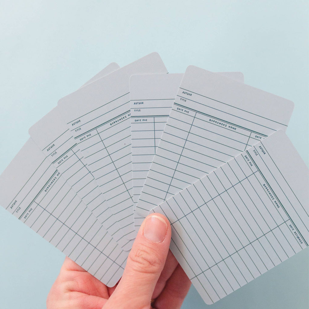 A hand holds a fanned stack of Sky Blue Library Card Deck Of Playing Cards, each featuring lines and headings for author, title, date due, and borrower's name, set against a light blue background. Ships in approximately one week.