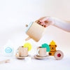 A hand pours from a Wooden Tea Set into a cup, surrounded by more wooden cups, saucers, a milk carton toy, honey bear figure, lemon slice, Matcha Tea bags, sugar cubes, and a biscuit with a red center—all in an imaginative play space.