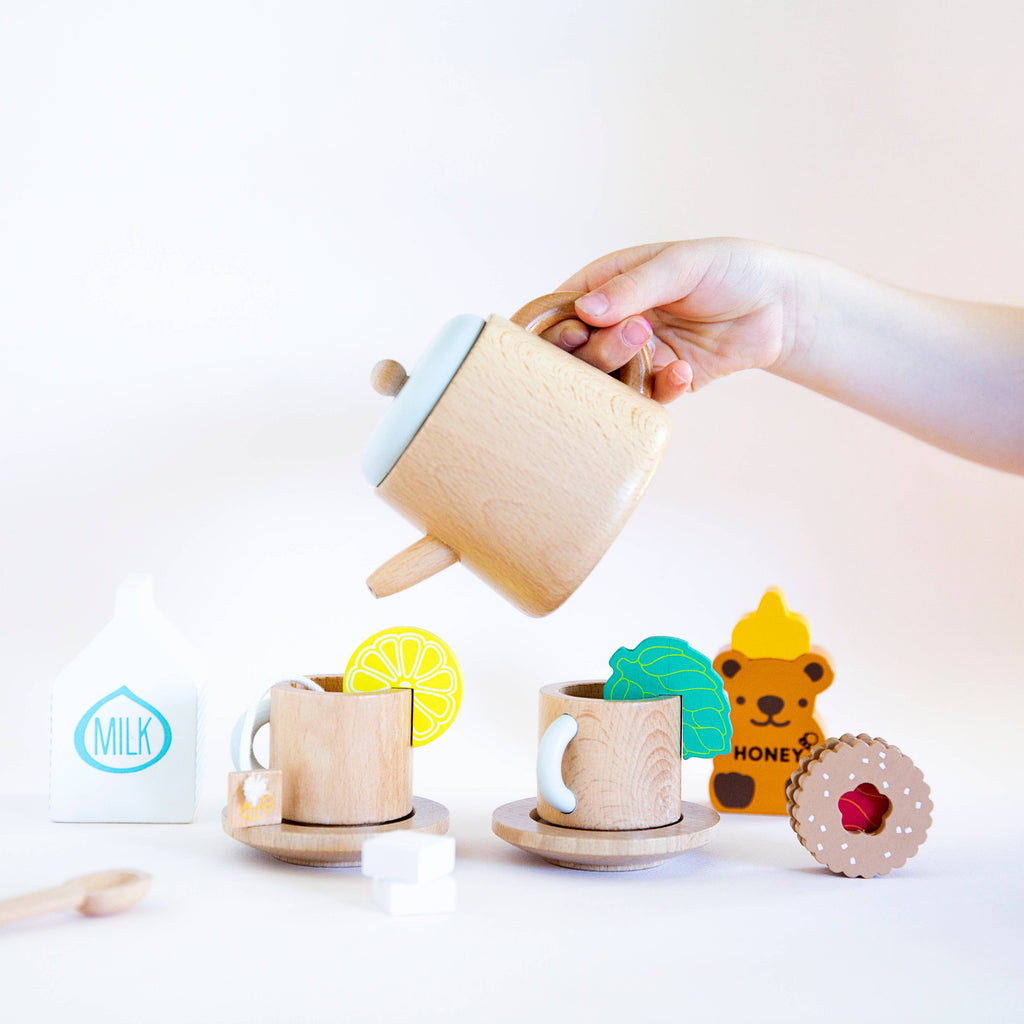 A hand pours from a Wooden Tea Set into a cup, surrounded by more wooden cups, saucers, a milk carton toy, honey bear figure, lemon slice, Matcha Tea bags, sugar cubes, and a biscuit with a red center—all in an imaginative play space.