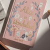 A pink hardcover "Alice in Wonderland | Collector's Edition" lies on a sunlit surface, decorated with flowers, playing cards, and illustrations of Alice and the White Rabbit.