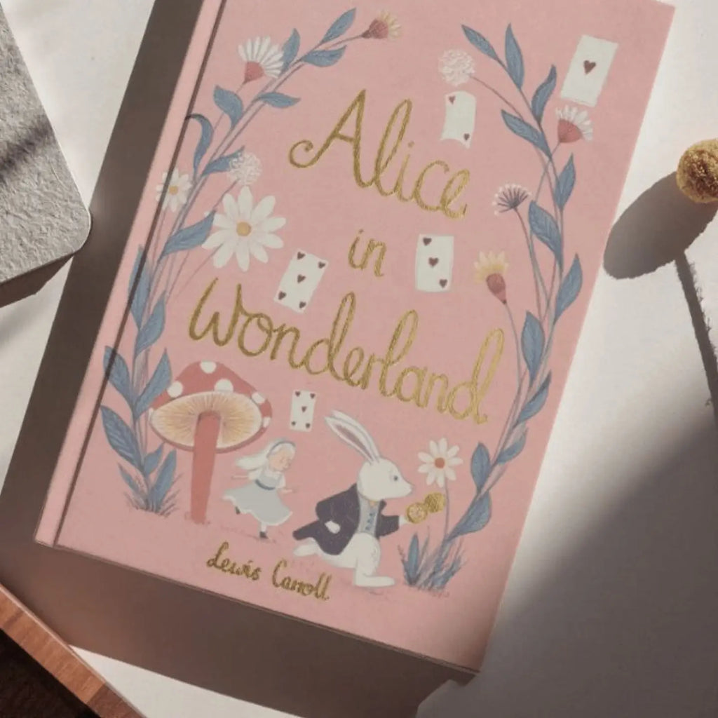 A pink hardcover "Alice in Wonderland | Collector's Edition" lies on a sunlit surface, decorated with flowers, playing cards, and illustrations of Alice and the White Rabbit.