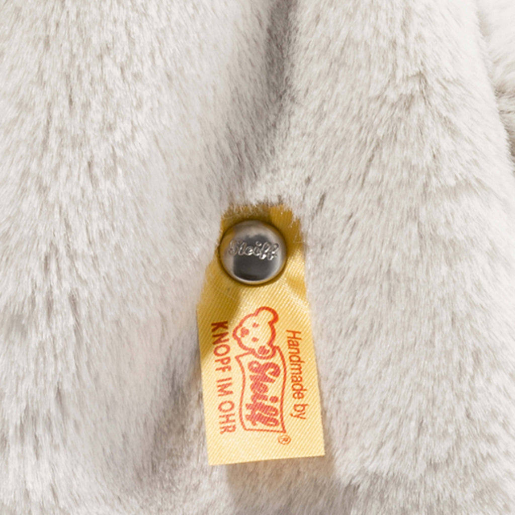 Close-up of a yellow Steiff tag and metal button attached to soft, light gray plush fabric, showing the Steiff Hoppie Bunny Stuffed Animal is handmade by Steiff.