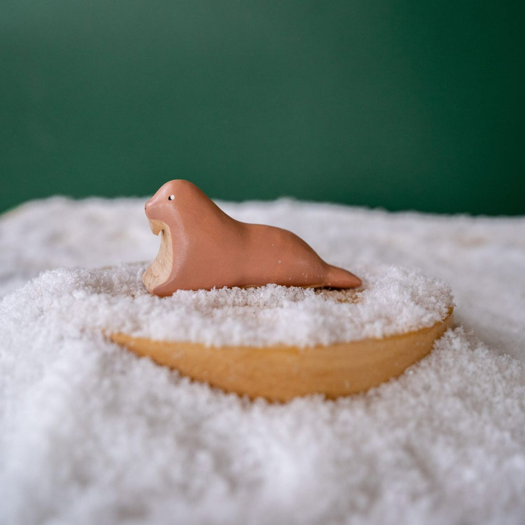 Handcrafted wooden walrus on a snowy surface, set on a round base. Painted with child-safe paint, it highlights natural beauty against a solid green background.