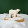 The Handmade Wooden Polar Bear is a charming small figurine crafted from natural wood, featuring child-safe paint. It stands on a wooden platform surrounded by artificial snow against a solid green background.