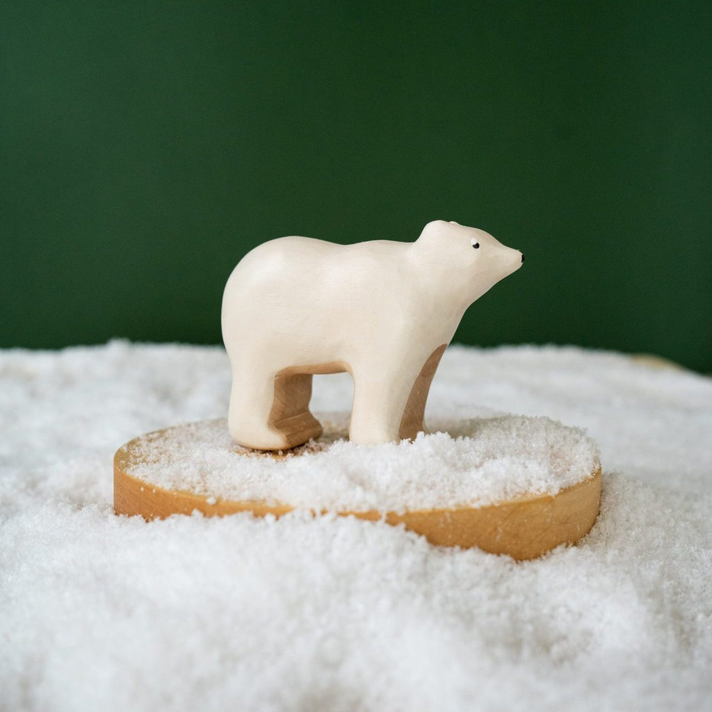 The Handmade Wooden Polar Bear is a charming small figurine crafted from natural wood, featuring child-safe paint. It stands on a wooden platform surrounded by artificial snow against a solid green background.