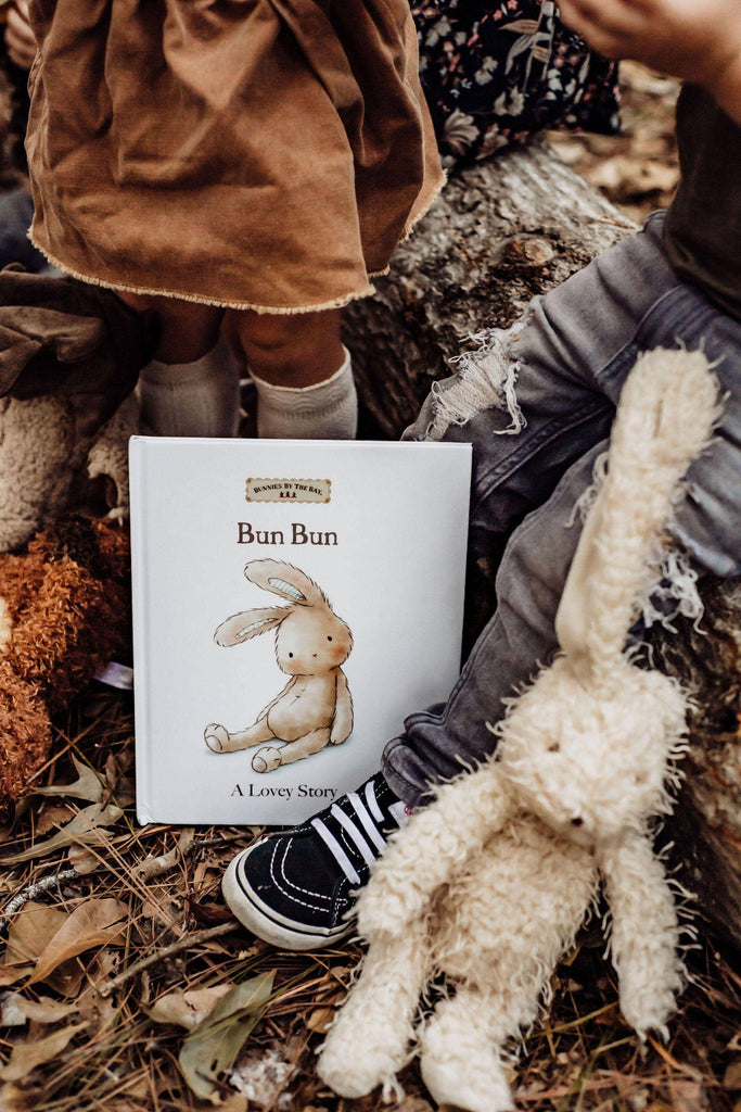 Children sit beside "Bun Bun: A Lovey Story" while a Harey the Bunny plush, with embroidered features, rests on their legs. Surrounded by leaves on the forest floor, the scene exudes a cozy storytelling ambiance with a vintage touch.