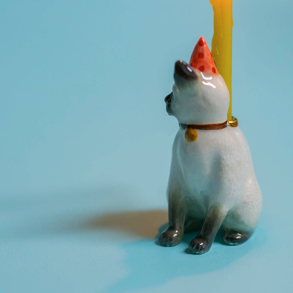 The Siamese Cat Cake Topper, a porcelain figurine with a birthday hat and yellow candle, sits against a light blue background, offering a lifelike and whimsical touch to cat-themed parties.