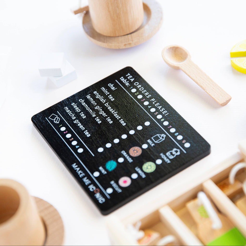 A delightful Wooden Tea Set on a white surface features a black menu card with organic Matcha Tea options, accompanied by a small wooden spoon, cup, and tea items that invite imaginative play.