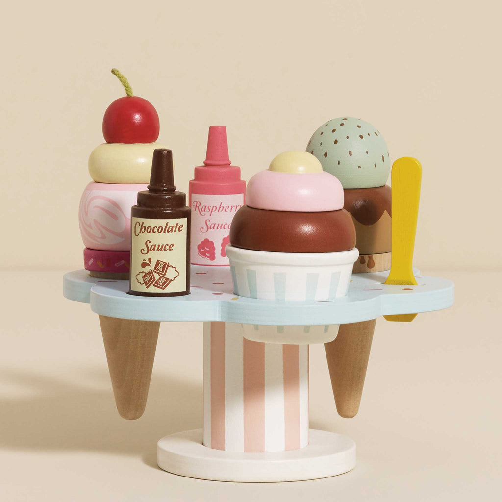 The Wooden Ice Cream Stand & Toppings features pastel-colored scoops, chocolate and raspberry sauce bottles, a cherry for topping, and a yellow spoon for delightful pretend serving. Perfect for role play fun.
