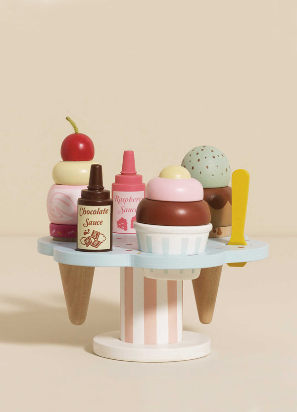 The Wooden Ice Cream Stand & Toppings is a whimsical toy with a pastel blue base, colorful scoops, cherry on top, sauce bottles, and a yellow spoon. It has a playful and smooth cartoonish design that’s ideal for role play fun.