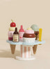 The Wooden Ice Cream Stand & Toppings is a whimsical toy with a pastel blue base, colorful scoops, cherry on top, sauce bottles, and a yellow spoon. It has a playful and smooth cartoonish design that’s ideal for role play fun.