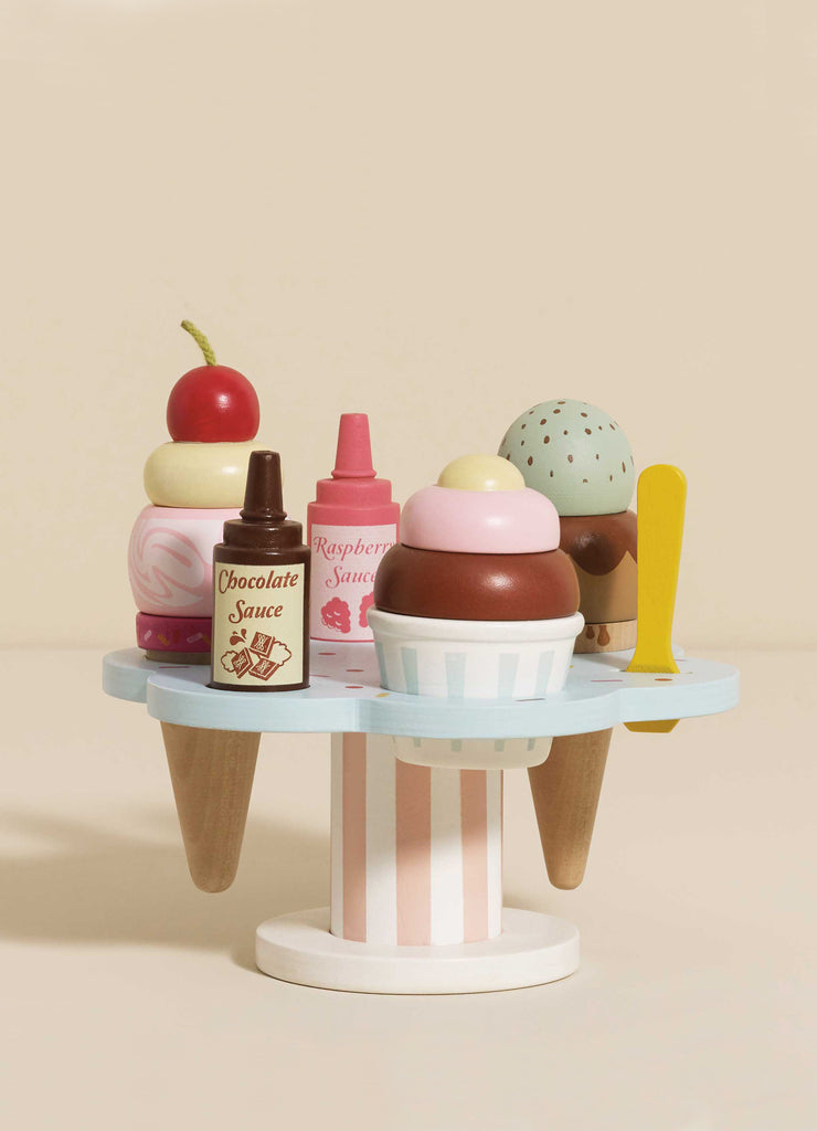 The Wooden Ice Cream Stand & Toppings is a whimsical toy with a pastel blue base, colorful scoops, cherry on top, sauce bottles, and a yellow spoon. It has a playful and smooth cartoonish design that’s ideal for role play fun.