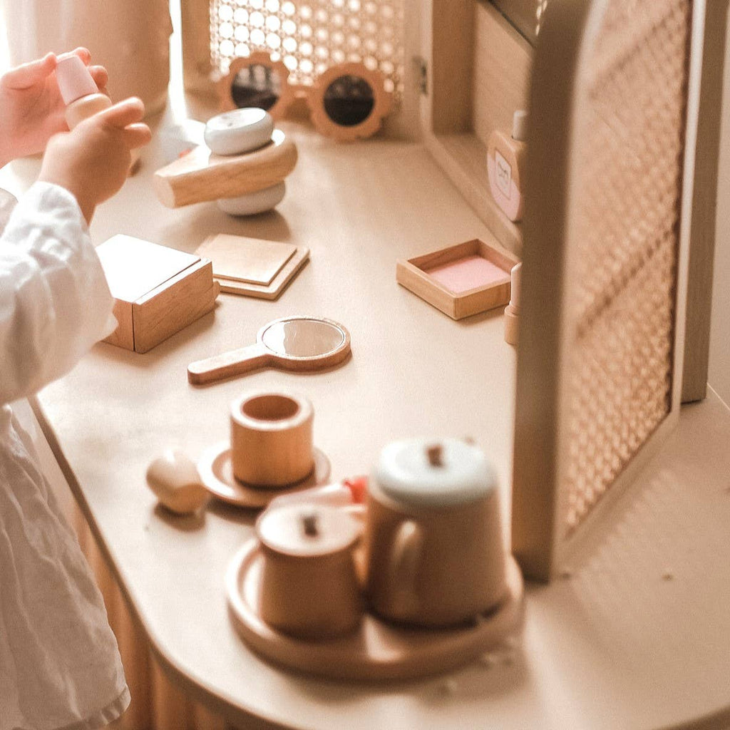 A child's hands interact with the Wooden Beauty Kit on a light surface. Painted with non-toxic paint, it includes pots, cups, a mirror, and various containers. A woven pattern and soft lighting enhance the toy's warm, cozy atmosphere.