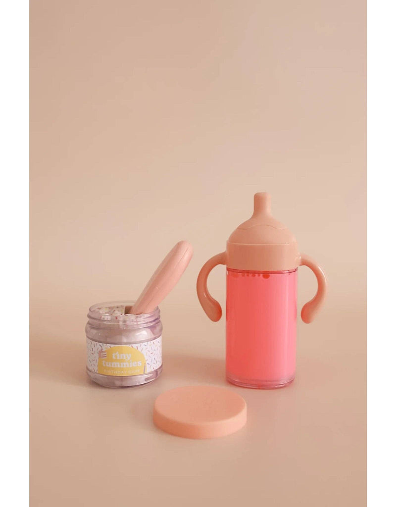 The My Tiny Birthday Set - Tiny Harlow includes a whimsical small jar called "tiny tummies" and a pink two-handled sippy cup, perfect for realistic play and delightful imaginary tea parties with birthday cake jars and strawberry milk, all set against a beige backdrop.