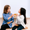 Two young girls enjoy a pretend tea party with the Wooden Tea Set. One wears a blue dress, the other a white sweater, as they face each other with small wooden cups and saucers, imagining almond chai, against a plain background.