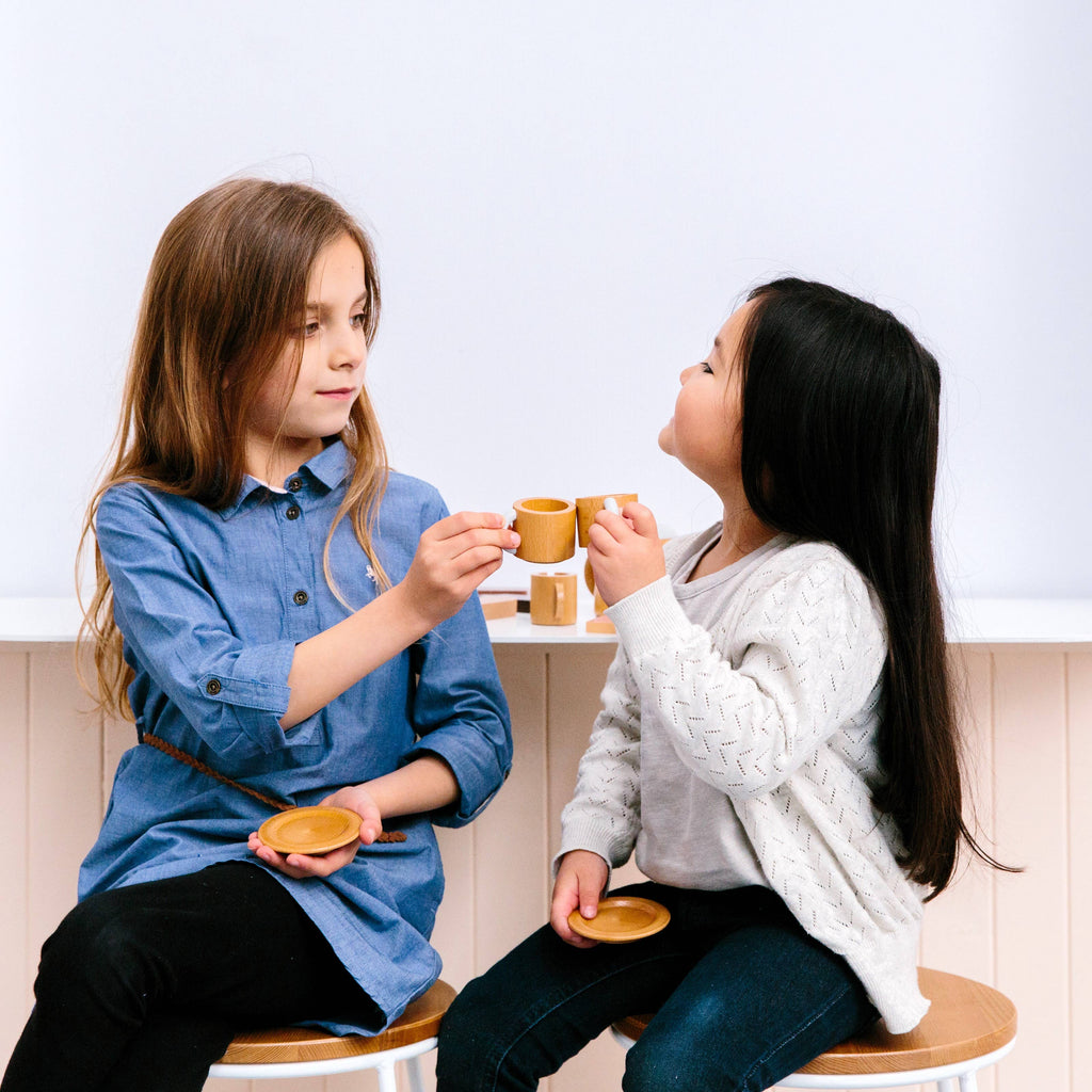 Two young girls enjoy a pretend tea party with the Wooden Tea Set. One wears a blue dress, the other a white sweater, as they face each other with small wooden cups and saucers, imagining almond chai, against a plain background.