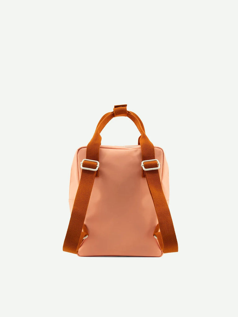 The Suzy Blush backpack from Sticky Lemon's Envelope Collection is salmon pink, made from waterproof nylon, features adjustable brown straps with metal buckles, and is shown from the back on an off-white background.