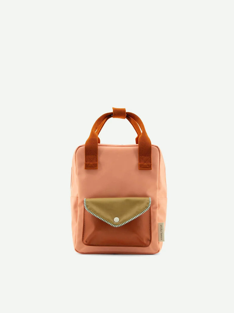 The Suzy Blush backpack from Sticky Lemon's Envelope Collection is small, square-shaped, and orange, made of waterproof nylon with a brown front pocket secured by a gold button. It has two short orange handles and a beige side label against a plain white background.