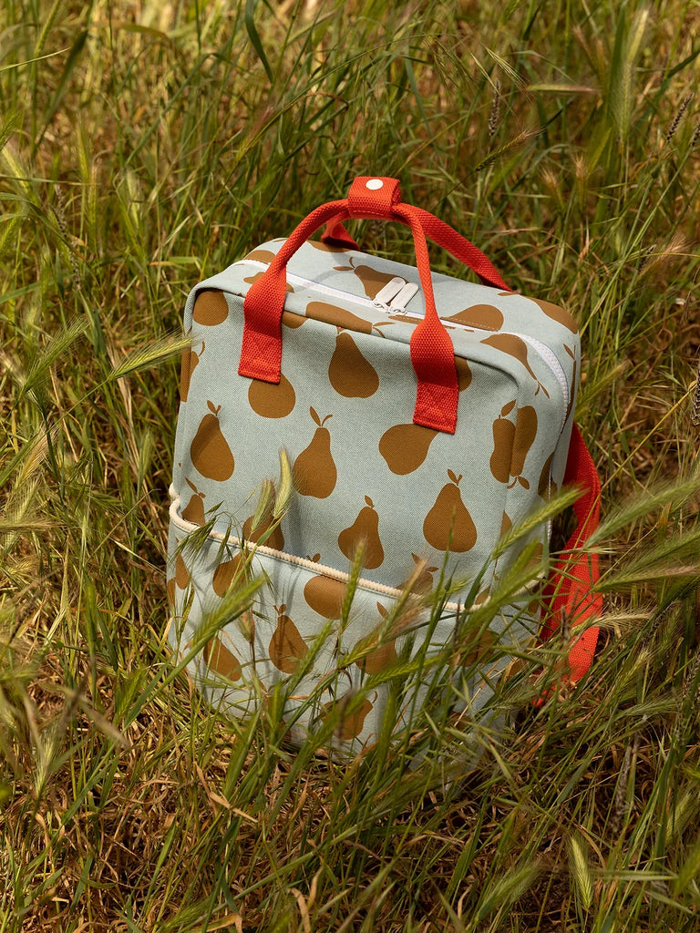 A Sticky Lemon Backpack Large with a pear print and red handles sitting in tall green grass, featuring a reliable YKK zipper for secure closure.