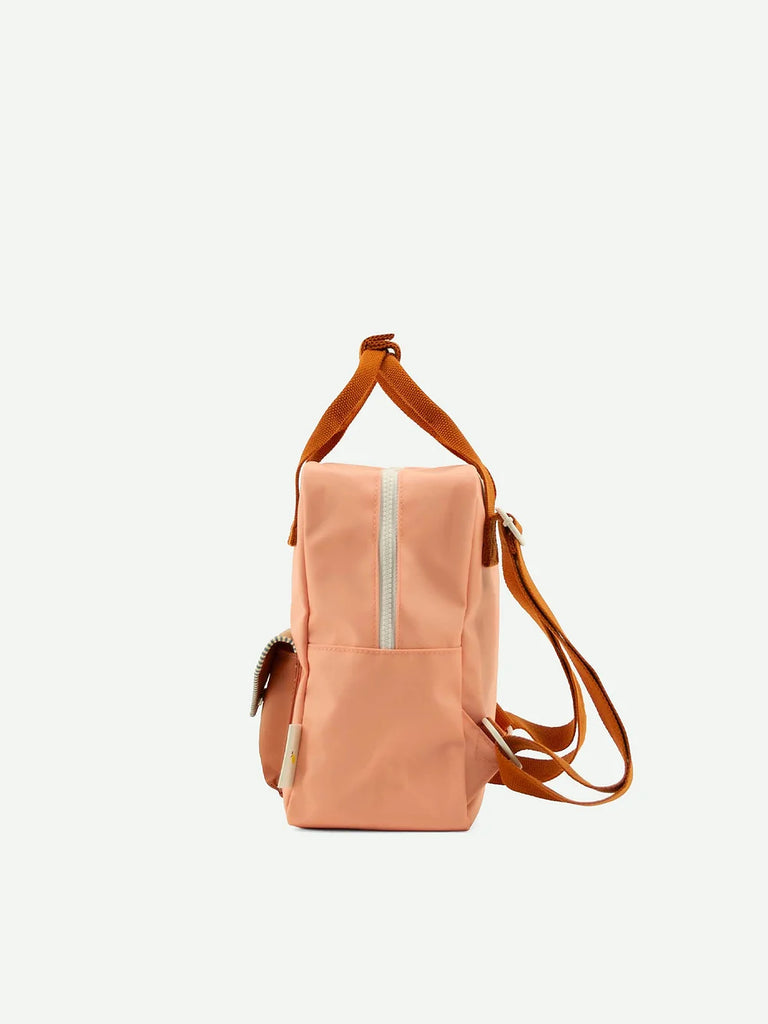 The Suzy Blush from Sticky Lemon's Envelope Collection is a peach-colored backpack made from recycled PET bottles, featuring brown straps, a front zipper pocket, and a shoulder handle. The plain light background accentuates its design.