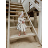 A young child in a white headband and dress sits barefoot on wooden stairs outside, holding a Senger Naturwelt Stuffed Animal - White Bunny and looking to the side. Sand is visible below the stairs.