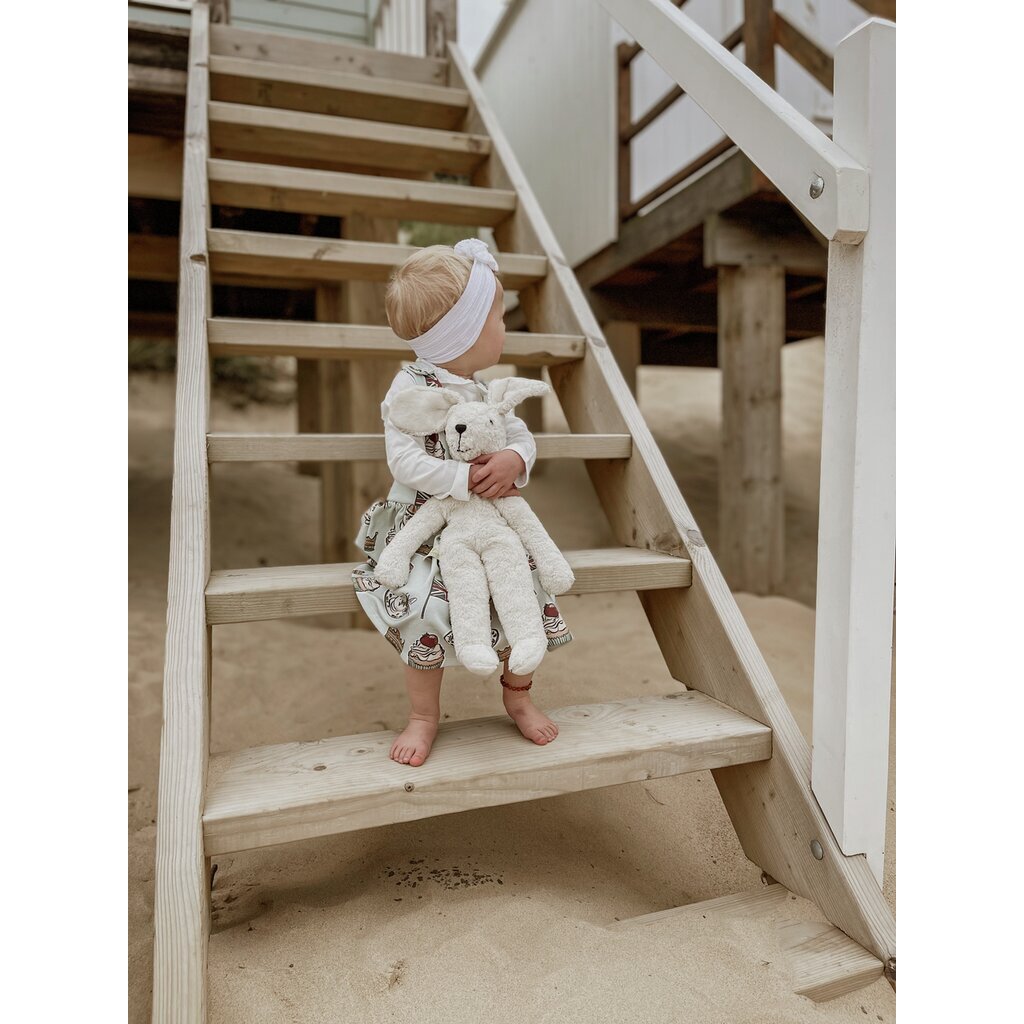A young child in a white headband and dress sits barefoot on wooden stairs outside, holding a Senger Naturwelt Stuffed Animal - White Bunny and looking to the side. Sand is visible below the stairs.