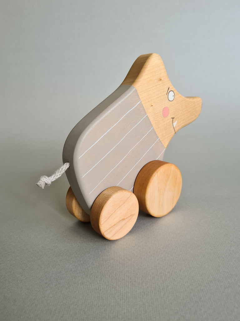 The Pull and Push Toy Little Wild Boar is a charming handcrafted toy featuring a wooden pig on wheels with a painted striped body, cheerful smile, and short string tail. Made from sustainable materials, it stands out beautifully against the plain gray background.