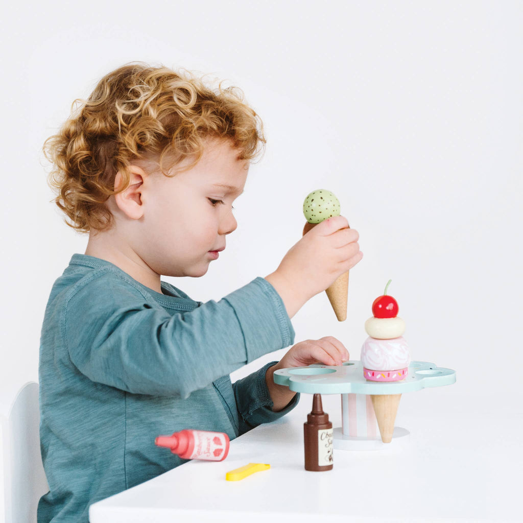 A curly-haired child enjoys role play with the Wooden Ice Cream Stand & Toppings, arranging a toy cone on the stand. Various colorful scoops and cones are displayed on the table against a plain white background, as they pretend to run their own gelato stand.