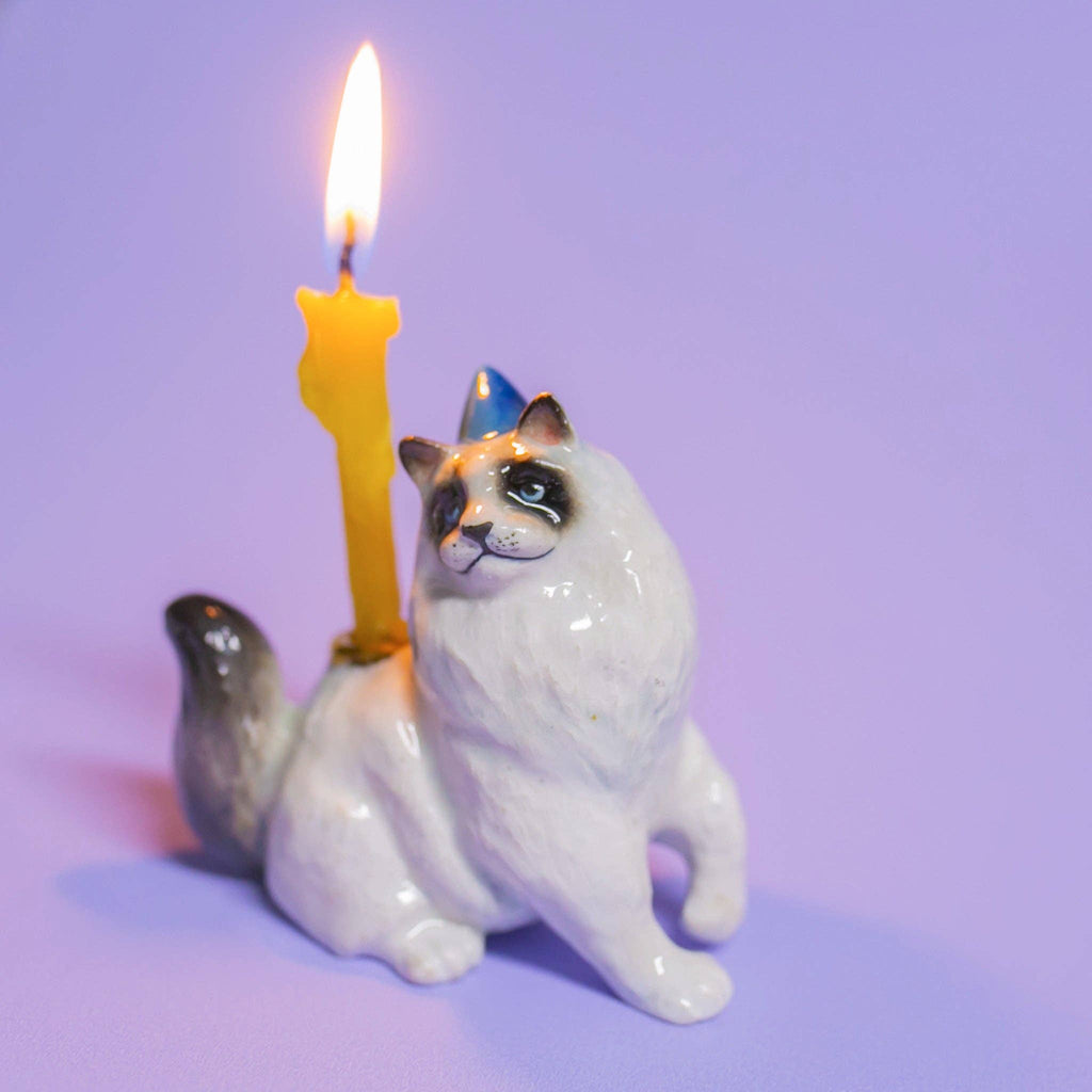 The Ragdoll Cat Cake Topper is a ceramic figurine with a glossy finish, featuring a small lit yellow candle on its back. It's ideal for cat-themed parties. The scene is set against a smooth, light purple surface.