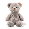 A fluffy, light gray Steiff Honey Teddy Bear, measuring 15 inches, features a brown nose and black eyes. It has a small yellow tag on its left ear identifying it as part of the Steiff plush toys collection and sits upright against a white background. This adorable bear is also machine washable for easy care.