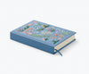 The Menagerie Garden Embroidered Journal is a blue hardcover with colorful floral and leaf embroidery on the cover, shown closed with a matching blue ribbon bookmark.