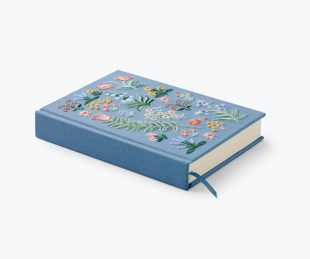 The Menagerie Garden Embroidered Journal is a blue hardcover with colorful floral and leaf embroidery on the cover, shown closed with a matching blue ribbon bookmark.