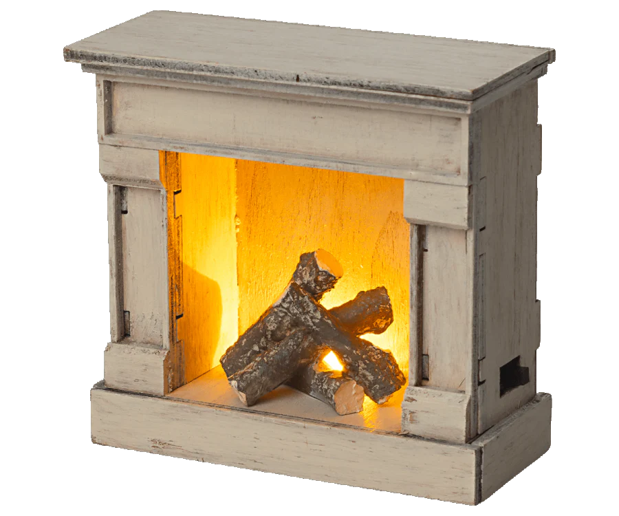 The Maileg Living Room Set includes a charming vintage-inspired fireplace model made of rustic wood, complete with faux logs and an internal glowing light for a cozy ambiance.