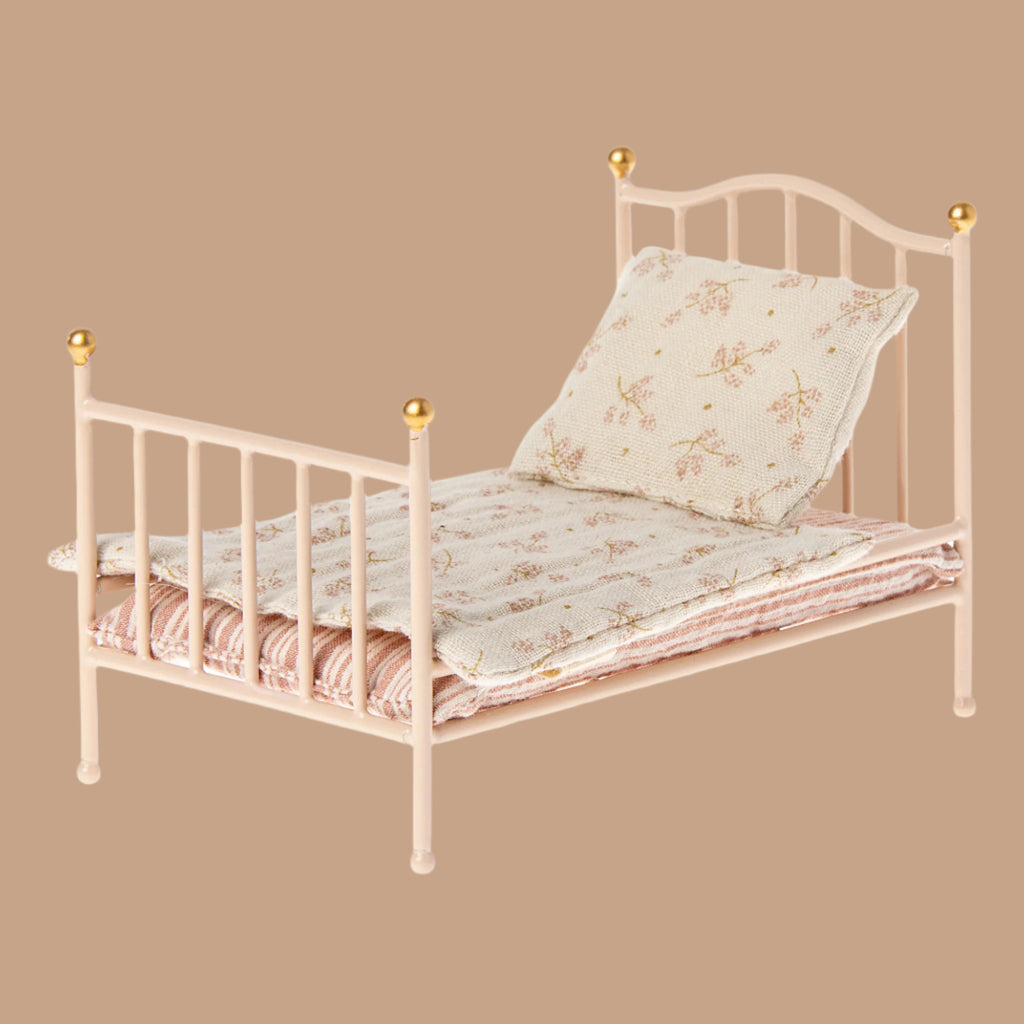 A Maileg Little Sister Bedtime Set in light pink, featuring gold-tipped posts, floral bedding, and a pillow is displayed against a plain beige background.