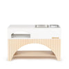 A modern white and light wood pet feeding station by Milton & Goose features two metal food bowls embedded in the countertop. Below, there is a shelf and a small storage cubby. The station has vertical wooden slats and an arched cutout at the base, echoing the stylish design of their Milton & Goose Mud Kitchen (Made in USA).