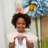 A curly-haired girl in a floral dress wears an Embellished Gingham Bunny Headband and holds a bunny-eared basket, with a large blue and yellow organza flower decorating the background.