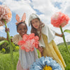 In a green field under a blue sky with fluffy clouds, two children play. One wears an Embellished Gingham Bunny Headband and holds pink flowers, while the other dons a cape and hat with a single bloom. Oversized paper flowers and delicate organza blooms create a whimsical backdrop.