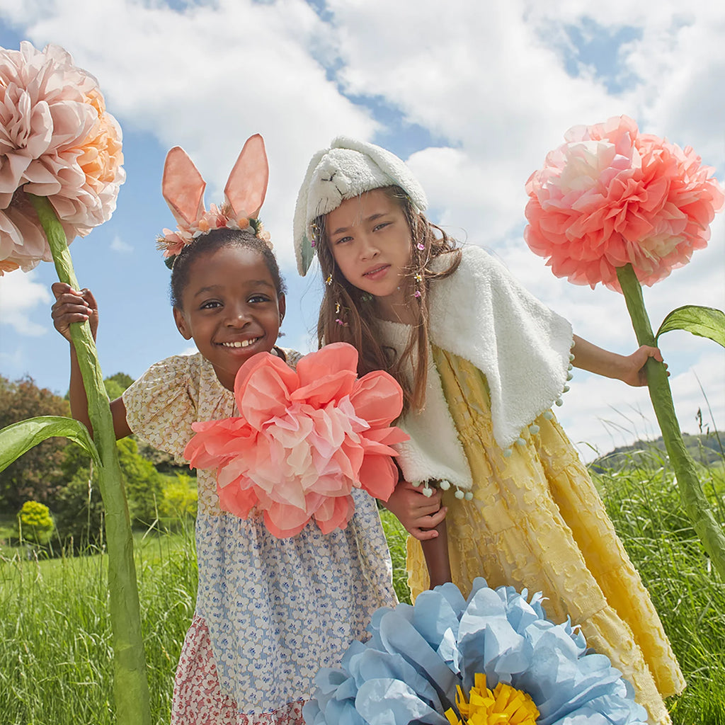 In a green field under a blue sky with fluffy clouds, two children play. One wears an Embellished Gingham Bunny Headband and holds pink flowers, while the other dons a cape and hat with a single bloom. Oversized paper flowers and delicate organza blooms create a whimsical backdrop.