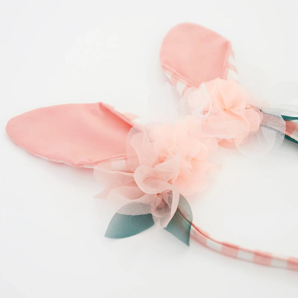 The Embellished Gingham Bunny Headband features soft pink rabbit ears adorned with peach tulle and green leaves on a white background, with organza flowers adding a whimsical, delicate touch.