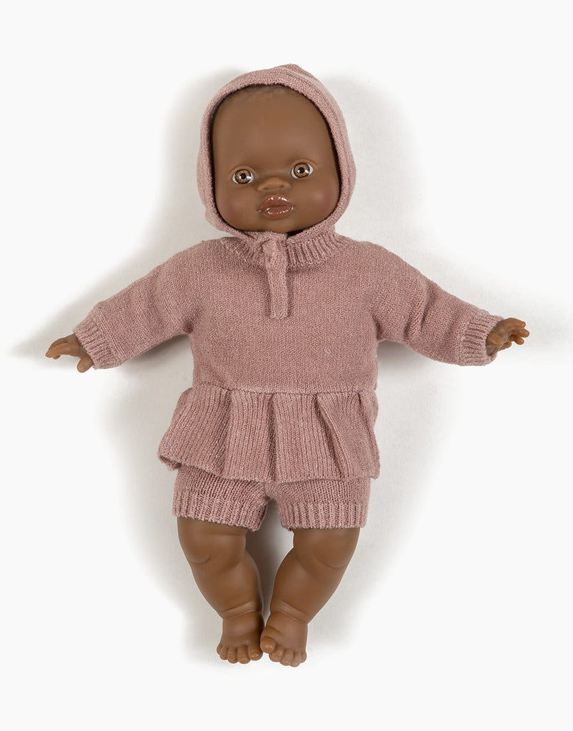 A Minikane Babies doll with medium brown skin is dressed in a Minikane Doll Clothing | Élie Tea Pink Knit Bonnet and matching shorts. The doll's eyes and lips are painted, showcasing the intricate details of the doll clothing, and it is positioned against a plain white background.