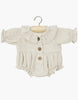 A beige, long-sleeved romper with a ruffled collar and cuffs, designed for the Minikane Sitting Doll (13"), hangs on a wooden hanger. It features three wooden buttons down the front and a gathered waist, complementing Charlie's anatomically correct form charmingly.