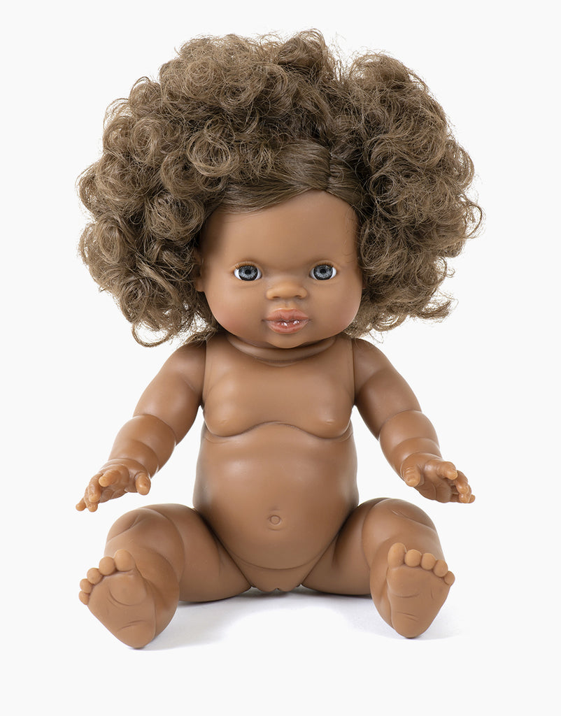 The Minikane Sitting Doll (13") - Charlie, featuring curly hair and brown skin, sits upright in detailed realism, proffering a gentle expression with blue eyes. This anatomically correct doll is unclothed and lifelike, emitting a delicate vanilla scent against a white backdrop.