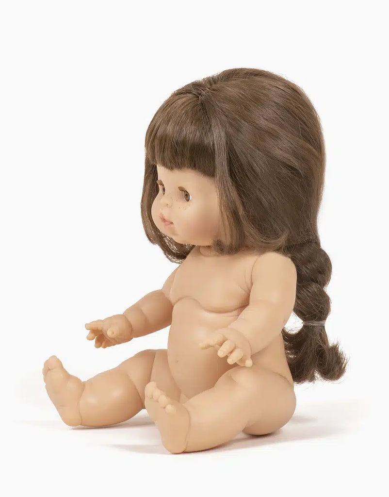 The Minikane Baby Doll (13") - Valentine With Outfit, featuring long brown hair and anatomically correct features, sits nude against a white background, facing left with slightly extended movable arms and bent knees in a lifelike pose.
