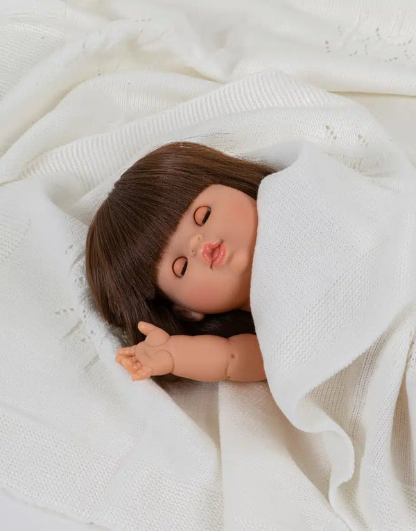 A Minikane Baby Doll with Sleeping Eyes (13") - Chloe, featuring brown hair and closed eyes, rests on a patterned white blanket with a raised left arm, radiating peace and a natural vanilla scent.