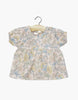The Minikane Doll Clothing Faustine Dress with Balloon Sleeves Célia showcases a pastel floral pattern in blue, yellow, and pink on cream. Designed for Minikane Babies or 11" dolls, it features a round neckline with gathering detail and hangs elegantly on a wooden hanger against a white background.