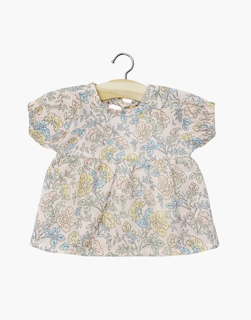 The Minikane Doll Clothing Faustine Dress with Balloon Sleeves Célia showcases a pastel floral pattern in blue, yellow, and pink on cream. Designed for Minikane Babies or 11" dolls, it features a round neckline with gathering detail and hangs elegantly on a wooden hanger against a white background.