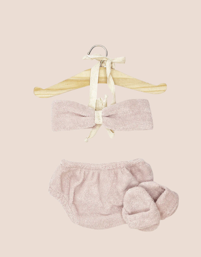 A soft pink baby outfit, perfect for the Minikane Standing Doll (14") like Jade With Clothing & Sunglasses, includes a headband, bloomers, and booties on a wooden hanger against a beige backdrop. The knitted texture and bow detail on the headband enhance each doll's vanilla scent appeal.