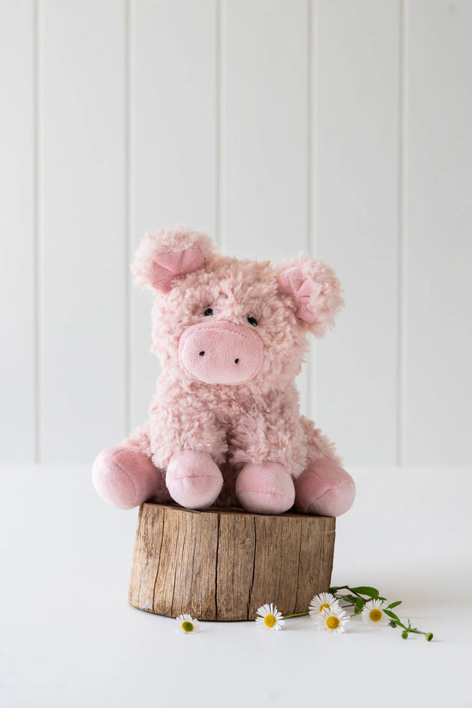 Poppy The Pig Stuffed Animal is perched on a small wooden stump against a white background, echoing *Animal Farm*, with a few small white flowers scattered nearby.